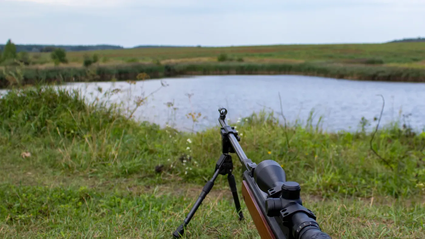 Gevär uppställt på bipod med sikte mot sjö och fält